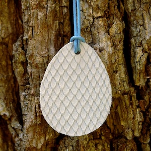 Könnte beinhalten: Ein cremefarbener, tropfenförmiger Anhänger mit einem strukturierten, schuppenartigen Muster, der an einer hellblauen Kordel hängt. Der Anhänger hängt vor einem Hintergrund aus rauer, brauner Baumrinde und erzeugt einen natürlichen Kontrast.