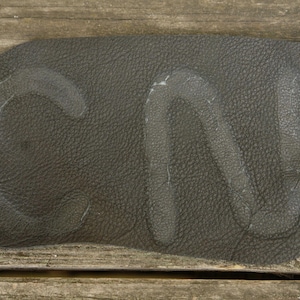 Könnte beinhalten: Ein dunkelgraues Lederpatch mit den Buchstaben "CN" auf der Oberfläche. Das Leder hat eine strukturierte Optik und liegt auf einem verwitterten Holzgrund.