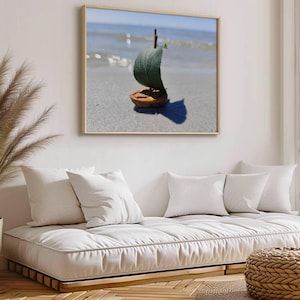 May include: A framed photograph of a small sailboat made from a walnut shell and a green leaf, floating on a sandy beach with blue ocean waves in the background.