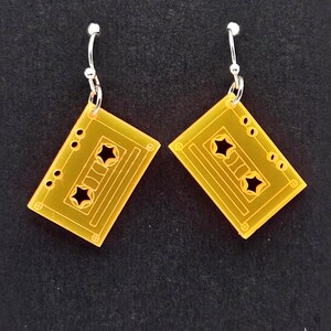 May include: A pair of translucent yellow acrylic earrings shaped like rectangular cassette tapes. Each earring features two star-shaped cutouts in the center and silver-toned metal fishhook ear wires for attachment to the earlobes.