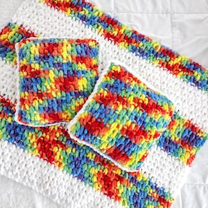 May include: A white crocheted throw blanket with rainbow stripes and two rainbow crocheted throw pillows.