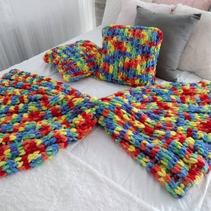 Könnte beinhalten: Eine regenbogenfarbene, grob gestrickte Wolldecke und zwei passende Kissen. Die Decke ist in der Mitte gefaltet und die Kissen liegen darauf. Decke und Kissen liegen auf einem weißen Bett.