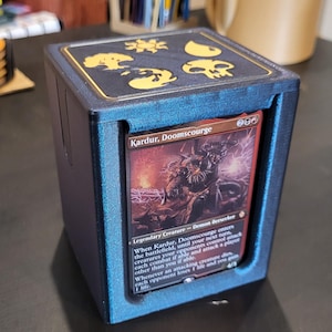 Könnte beinhalten: Eine blaue und goldene Kartenaufbewahrungsbox mit einer Karte mit dem Text "Kardur, Doomscourge". Die Box hat eine Oberseite mit goldenen Symbolen. Die Karte ist auf der Vorderseite der Box sichtbar.