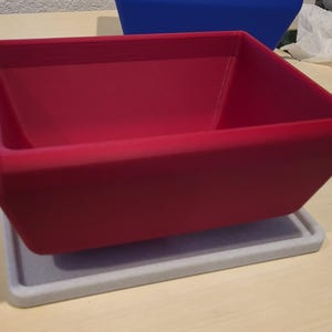 May include: A red square planter box with a matching grey rectangular base. The planter is a vibrant red colour and has a smooth, matte finish. The base is a light grey colour. The planter is sitting on a light-coloured surface.