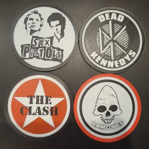 May include: Four round coasters featuring iconic punk rock band logos. The coasters include designs for Sex Pistols, Dead Kennedys, The Clash, and Ramones. Each coaster has a black border and a unique graphic.