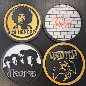 May include: Four round black coasters with gold trim. Each coaster features a different band logo: Jimi Hendrix, Pink Floyd, The Doors, and Led Zeppelin.