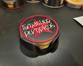 Puede incluir: Un juego de posavasos con la inscripción "Smashing Pumpkins" en escritura blanca sobre una estrella roja sobre fondo negro. Los posavasos se guardan en un soporte negro. Otros posavasos son visibles en el fondo.