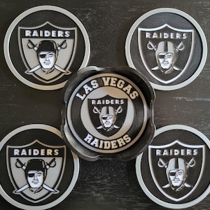 Op de afbeelding: Set van vijf ronde zwarte en zilveren onderzetters met het logo van de Las Vegas Raiders. Het logo toont een zwart-witte helm met gekruiste zwaarden en het gezicht van een speler.