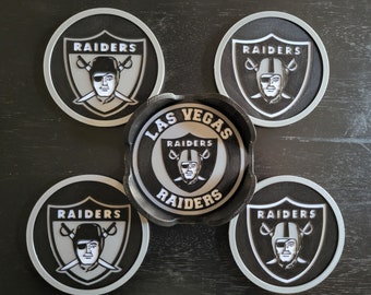 Puede incluir: Conjunto de cinco posavasos redondos negros y plateados con el logotipo de los Las Vegas Raiders. El logotipo presenta un casco negro y blanco con espadas cruzadas y la cara de un jugador.