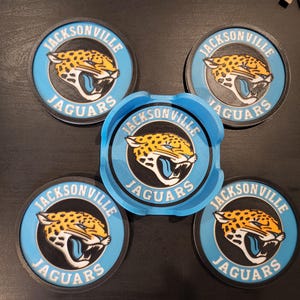 May include: A collection of Jacksonville Jaguars themed coasters and an ashtray. The coasters and ashtray are blue with a black border and feature the team's logo: a jaguar head in orange, black, and white, with the words "Jacksonville Jaguars".