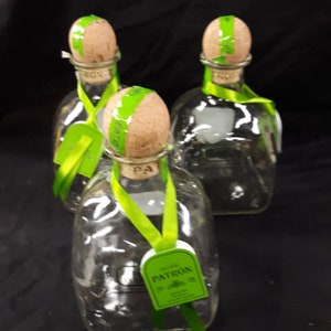 May include: Three clear glass bottles with cork tops and green ribbon accents. The bottles are labeled "Patron" and have a green and white label with a leaf design.