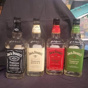 May include: Four glass bottles of Jack Daniel's Tennessee Whiskey. The bottles are arranged from left to right: Old No. 7, Tennessee Honey, Tennessee Fire, and Tennessee Apple. Each bottle has a different label design with a black, white, red, and green color scheme.