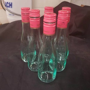 May include: Six clear glass bottles with pink screw tops. The bottles are empty and have a textured surface. The bottles are arranged in a circle on a black surface.