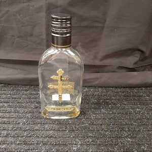 May include: A clear glass bottle with a black cap and a gold label. The label features a stylized design of a cross with a sword. The bottle is labeled "Dusse" and is sitting on a gray textured surface.