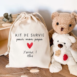 Peut inclure: Un sac en coton beige avec le texte "KIT DE SURVIE pour mon papa" et "J'arrive ! Alba" imprimé en noir, avec un cœur rouge. Deux ours en peluche, un marron et un blanc, sont à côté du sac.