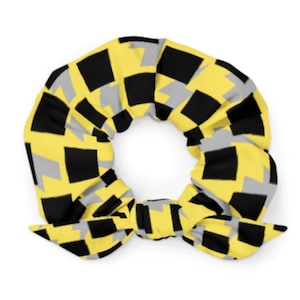 May include: A yellow, black, and gray patterned scrunchie. The scrunchie features a repeating geometric design of squares in yellow, black, and gray. The scrunchie is tied in a bow at the bottom.