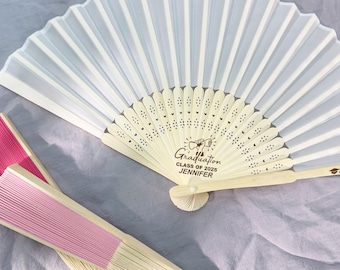 May include: White and pink folding fans with wooden handles. The white fan has the text "Graduation Class of 2025 Jennifer". The pink fan is folded and has a light pink pleated design. Graduation party favors.