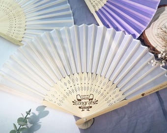 May include: White and purple hand fans with wooden handles. The white fan has a laser-cut design and the text "Class of 2025 Congrats! Michelle".