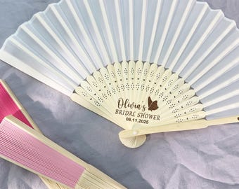 May include: White and pink folding fans with wooden handles. The white fan is open, displaying the text "Olivia's Bridal Shower 08.11.2025" with a butterfly graphic. The pink fans are closed.