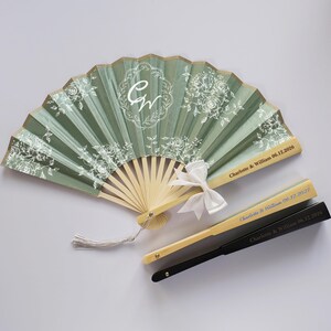 May include: A sage green folding hand fan with white floral patterns, a central monogram with the letters CW, and a white ribbon bow. Two closed fans are positioned below, one light wood with blue text and one black with white text.