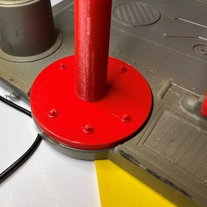 Può includere: Una base di plastica rossa con un palo di plastica blu e bianco attaccato. La base ha sei piccole viti attorno al bordo.
