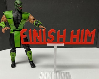 May include: A green and black action figure of a character from the Mortal Kombat video game series, standing in front of a red 3D printed sign that reads "FINISH HIM".