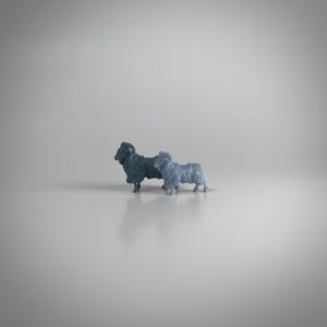 May include: Two small, detailed ram figurines in shades of blue-grey stand side-by-side. The rams have textured wool-like coats and curved horns, with a neutral background. These miniature sculptures could be used as decorative items or collectibles.