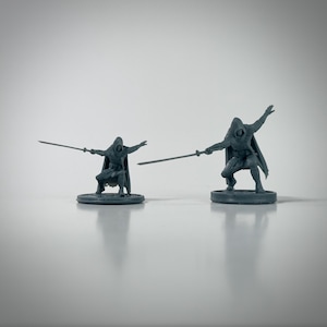 May include: Two gray miniature figures of hooded warriors in action poses, each holding a sword. The figures are on circular bases, with one figure in a kneeling position and the other in a dynamic stance.