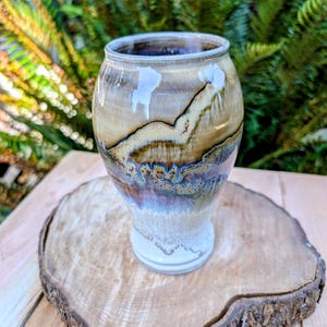 May include: A ceramic tumbler with a rounded body and a tapered base, featuring a multi-colored glaze with layers of cream, tan, brown, and blue. The surface displays a drip effect where the colors blend and overlap, creating a textured appearance.