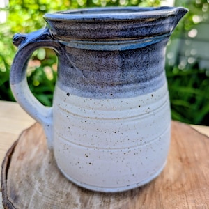 May include: A ceramic pitcher featuring a two-tone glaze with a dark blue, textured upper section and a speckled off-white lower body. The vessel includes a curved handle, a wide pouring spout, and horizontal ridges encircling the base.