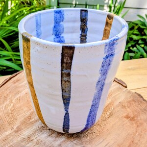 May include: A white ceramic tumbler featuring a series of vertical stripes in shades of blue, dark brown, and metallic gold. The vessel has a slightly tapered cylindrical shape with a textured, hand-thrown appearance and a smooth, glossy finish.