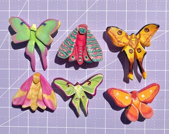 May include: Six colorful wooden butterfly pins with intricate details. The butterflies are painted in shades of pink, yellow, green, and blue.