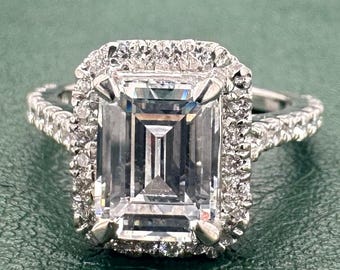 May include: A silver ring featuring an emerald-cut diamond, encircled by smaller diamonds. The ring is displayed on a green surface. The brand name "Jenu Jewel" is visible in the top right corner.