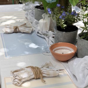 May include: A table setting with blue hydrangeas, candles, and place settings. The place mats are light blue and cream with a striped design. Napkins are tied with twine. Wine glasses and a small terracotta bowl are also on the table.