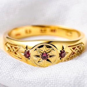 May include: A gold ring featuring a central starburst motif set with a small red gemstone, flanked by two additional smaller red gemstones on either side. The band displays an intricate engraved lattice pattern along the sides of the ring.