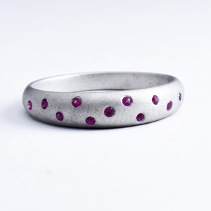 May include: A silver ring with a matte finish, featuring several small, round, ruby-coloured gemstones. The gemstones are evenly spaced around the band, creating a dotted pattern. The ring is set against a white background.