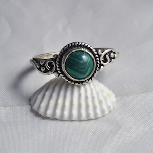 Könnte beinhalten: Ein Silberring mit einem runden, grünen Malachitstein, der in einer dekorativen Lünette gefasst ist. Der Ring hat ein verziertes Design mit Wirbelmustern auf dem Band. Der Ring wird auf einer weißen Muschelschale präsentiert.