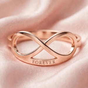 May include: A rose gold ring featuring an infinity symbol design. The band is smooth and polished with the word FOREVER engraved in capital letters along the bottom curve of the infinity loop, creating a continuous and symmetrical metal shape.