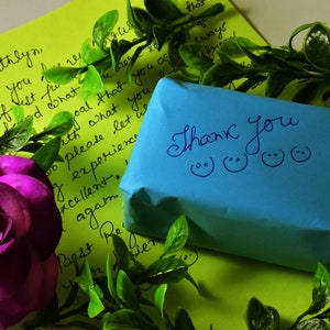May include: A vibrant composition featuring a purple rose, green foliage, and handwritten notes on lime green paper. A small blue package with the words "Thank You" and smiley faces is also present. The scene evokes a sense of gratitude and appreciation.