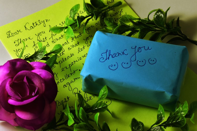 May include: A vibrant composition featuring a purple rose, green foliage, and handwritten notes on lime green paper. A small blue package with the words "Thank You" and smiley faces is also present. The scene evokes a sense of gratitude and appreciation.