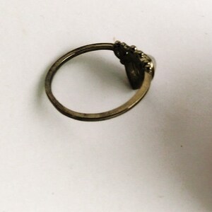 May include: A vintage-style ring with a tarnished gold finish. The ring features a decorative element with a dark, possibly stone-like, center. The band is thin and simple, with a slightly worn appearance, suggesting age or use.