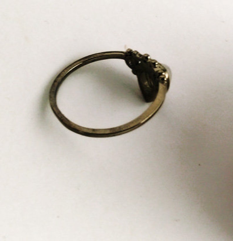 May include: A vintage-style ring with a tarnished gold finish. The ring features a decorative element with a dark, possibly stone-like, center. The band is thin and simple, with a slightly worn appearance, suggesting age or use.