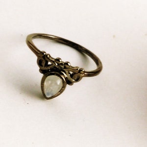 May include: A bronze-toned ring with an ornate design. The ring features a pear-shaped, iridescent gemstone at the center, set within a decorative filigree setting. The band has a delicate, curved design, suitable for everyday wear.