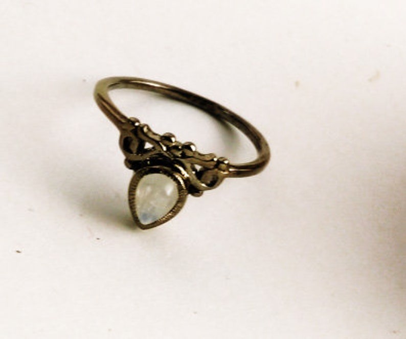 May include: A bronze-toned ring with an ornate design. The ring features a pear-shaped, iridescent gemstone at the center, set within a decorative filigree setting. The band has a delicate, curved design, suitable for everyday wear.