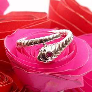 Op de afbeelding: Een zilverkleurige slangenring met een rode edelsteen oog, gewikkeld rond een roze roos. De ring heeft een gedetailleerd, getextureerd patroon en de kop van de slang is prominent aanwezig.