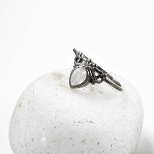 May include: A silver-toned ring with a pear-shaped, light-colored stone. The ring features an ornate, openwork design with a decorative top. The ring is displayed on a white, textured surface.