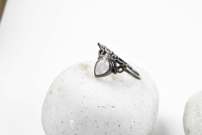 May include: A silver-toned ring with a pear-shaped, light-colored stone. The ring features an ornate, openwork design with a decorative top. The ring is displayed on a white, textured surface.