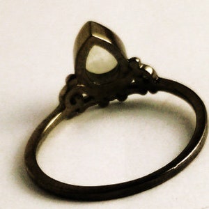 May include: A dark metal ring with an oval, light-colored gemstone. The ring has an ornate design around the gemstone and along the band. The ring is set against a light background.