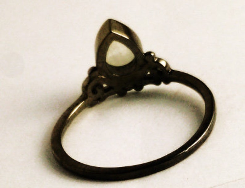 May include: A dark metal ring with an oval, light-colored gemstone. The ring has an ornate design around the gemstone and along the band. The ring is set against a light background.