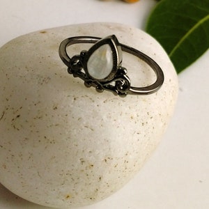 May include: A black metal ring with a pear-shaped, iridescent gemstone. The ring features ornate detailing around the gemstone and along the band. The ring is set on a white, textured surface, showcasing the jewelry's design and color.